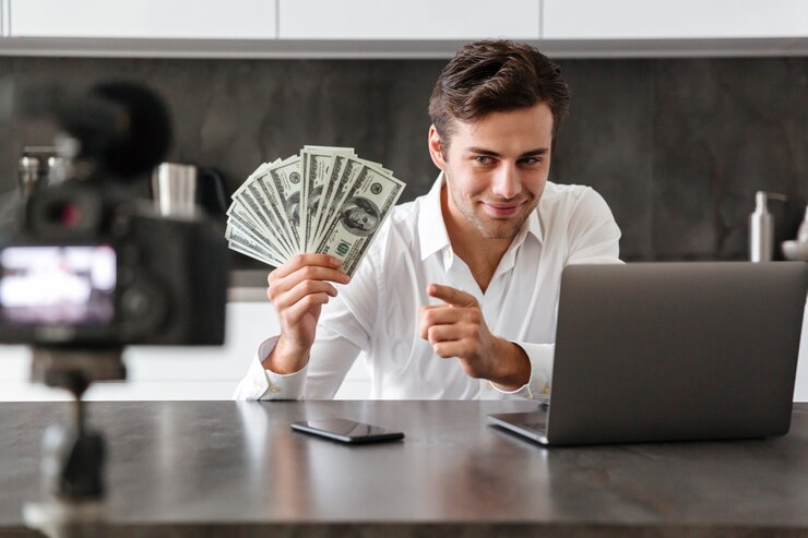 smiling young man filming his video blog episode about new tech devices while sitting kitchen table with laptop showing bunch money banknotes 171337 5530 1